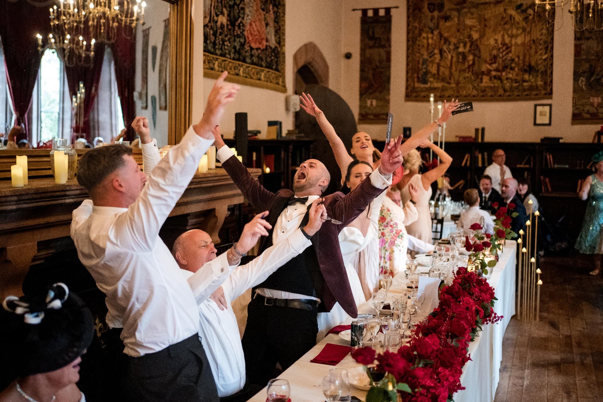 The bride and groom cheering with their wedding party at a long banquet table decorated with red roses in a candlelit stone hall