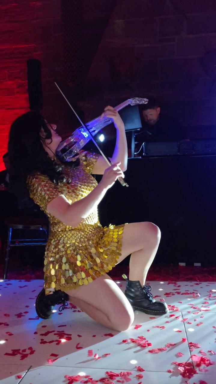 A live electric violinist kneeling on a starlit dancefloor, playing a glowing white electric violin while a DJ plays behind her