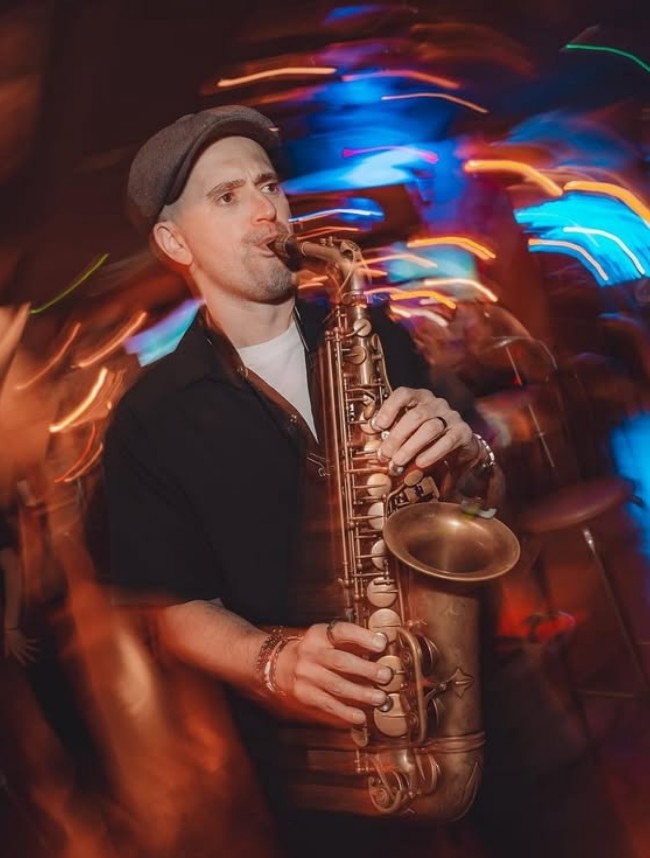 A live saxophonist in a flat cap playing a saxophone at an evening party, light trails swirling around him