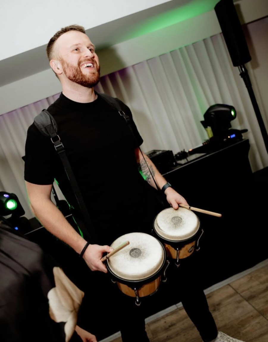 A smiling live percussionist playing bongos strapped to his waist at an evening party, green stage lights behind him