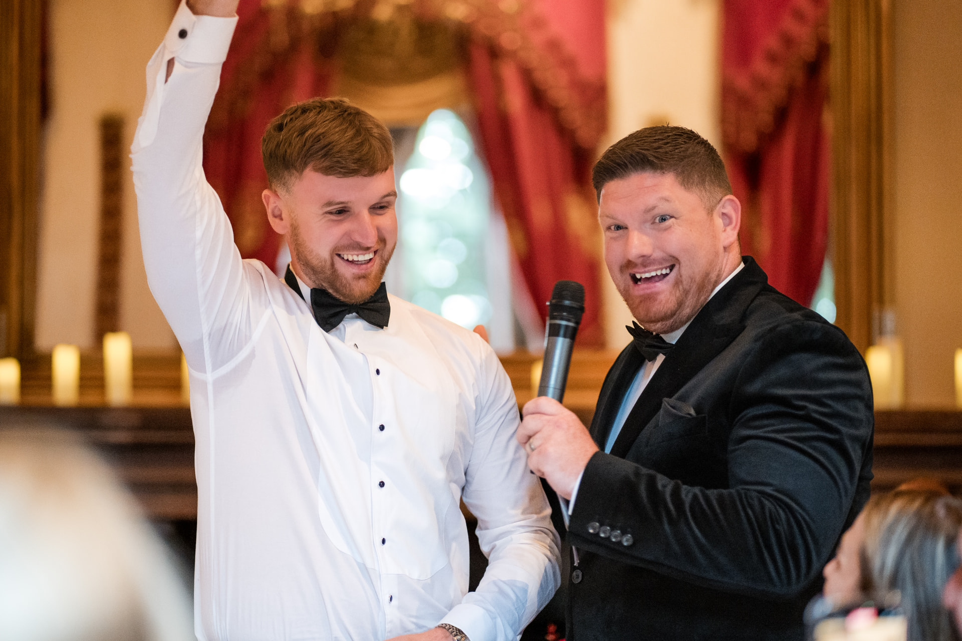 Jon Kane hosting with a microphone beside a groom with his arm raised in celebration